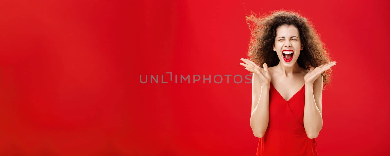 Woman releasing stress screaming out loud. Expressive and over-emotive attractive curly-haired woman in elegant red dress yelling with closed eyes and palms raised near face over studio background. Emotions concept