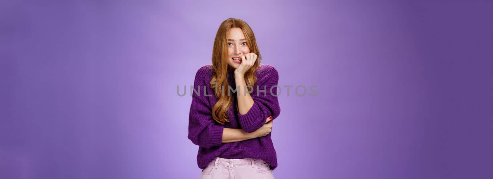 Portrait of nervous insecure cute redhead girl in purple sweater biting fingernails and stooping as feeling scared before exam or interview standing anxious and unsure over violet background by Benzoix