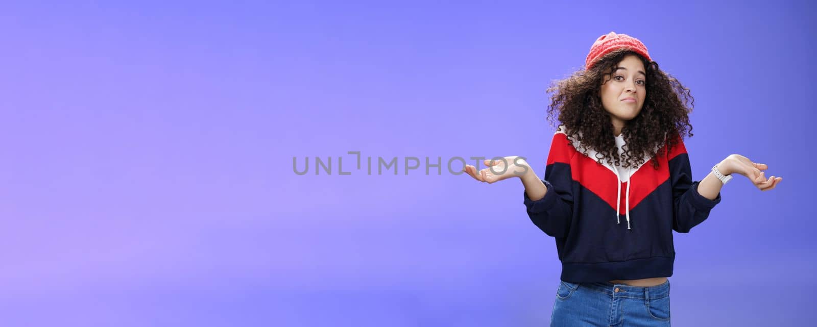 Upset girl cannot get clue what happened shrugging turning away as pouting feeling questioned and unsure what answer, standing clueless and puzzled in sweatshirt and stylish hat over blue background.