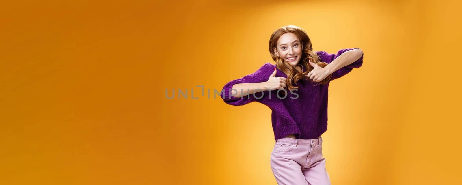 Optimistic and joyful bright ginger girl jumping happily and cheerful having fun showing thumbs up in like and approval giving positive feedback of super cool idea or concept over orange wall. Emotions concept