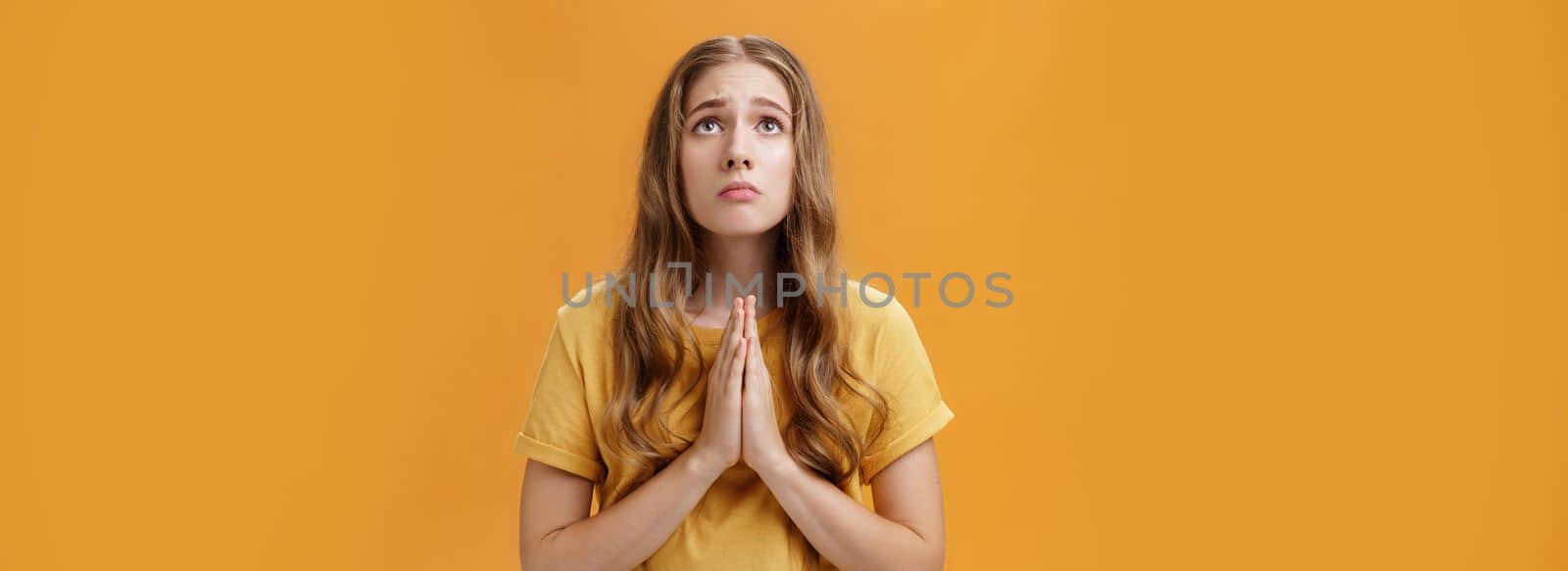 Hopeful uneasy and worried gloomy faithful girl in t-shirt holding hands in pray against chest looking up with sad look praying making wish for good well of family posing over orange wall by Benzoix