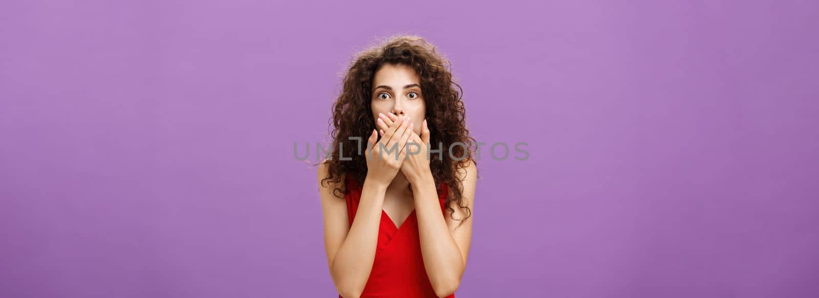 Cocnerned and shocked troubled adult woman with curly hairstyle in elegant evening red dress gasping from surprise and fear covering mouth with palms standing speechless over purple wall by Benzoix