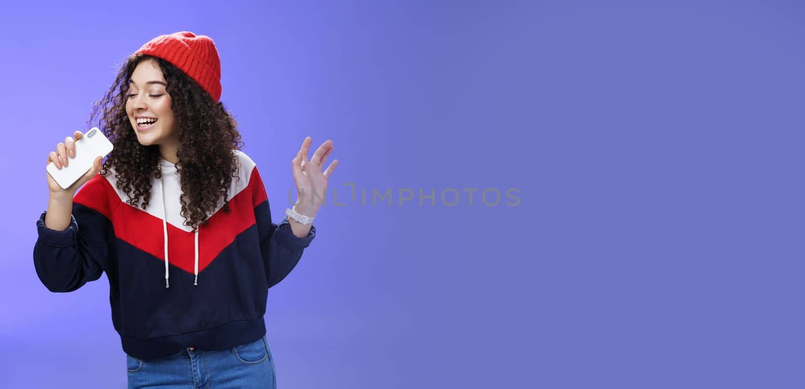 Attractive happy young woman with curly hair in hat singing enjoying perfect winter day singing along in smartphone, holding mobile phone like microphone, adore karaoke over blue background.