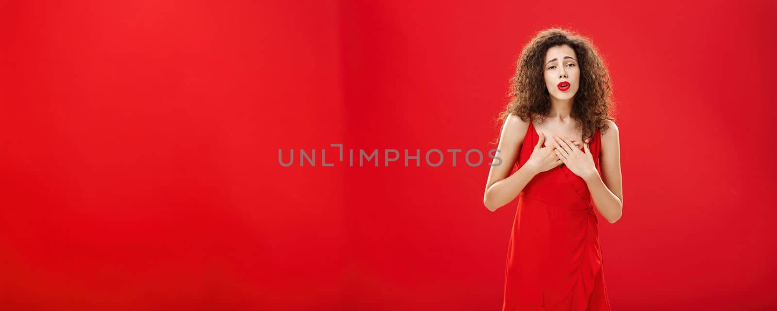 Portrait of touched elegant and stylish curly-haired adult. woman receiving compliments holding hands on chest in grateful and thankful pose hearing heartwarming speech in her honor over red background.