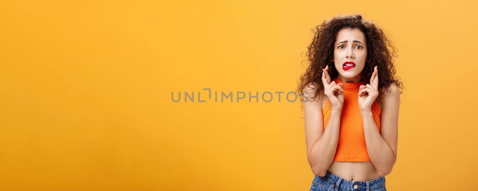 Worried and anxious silly caucasian female with curly hair in red lipstick and cropped top biting lower lip nervously looking concerned crossing fingers for good luck making wish over orange wall by Benzoix