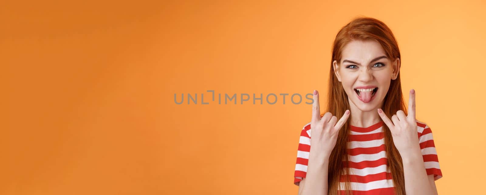 Lets rock-n-roll. Daring cheerful good-looking caucasian redhead woman, having fun, show heavy metal gestures, stick tongue and smile amused, enjoy music festival, stand orange background.