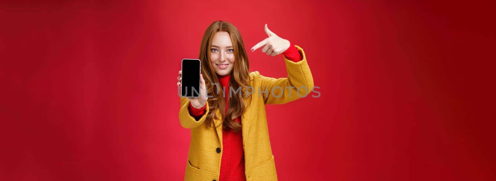 Pick this phone you never regret. Portrait of friendly-looking attractive and confident redhead glamour girlfriend in yellow coat showing smartphone pointing at mobile as smiling broadly at camera.