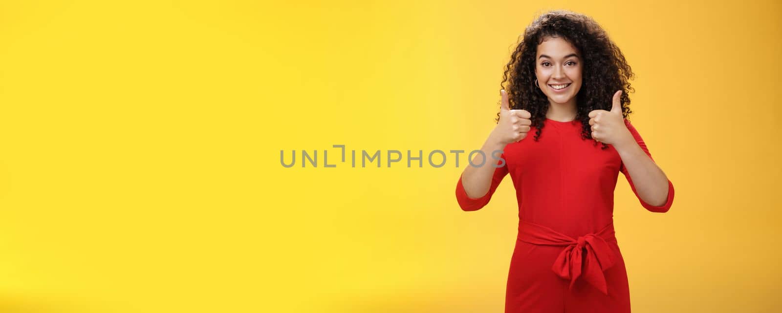 Excellent idea, nice job. Portrait of supportive delighted and happy charming female friend showing thumbs up as standing in red dress over yellow wall smiling giving positive reply, liking concept.
