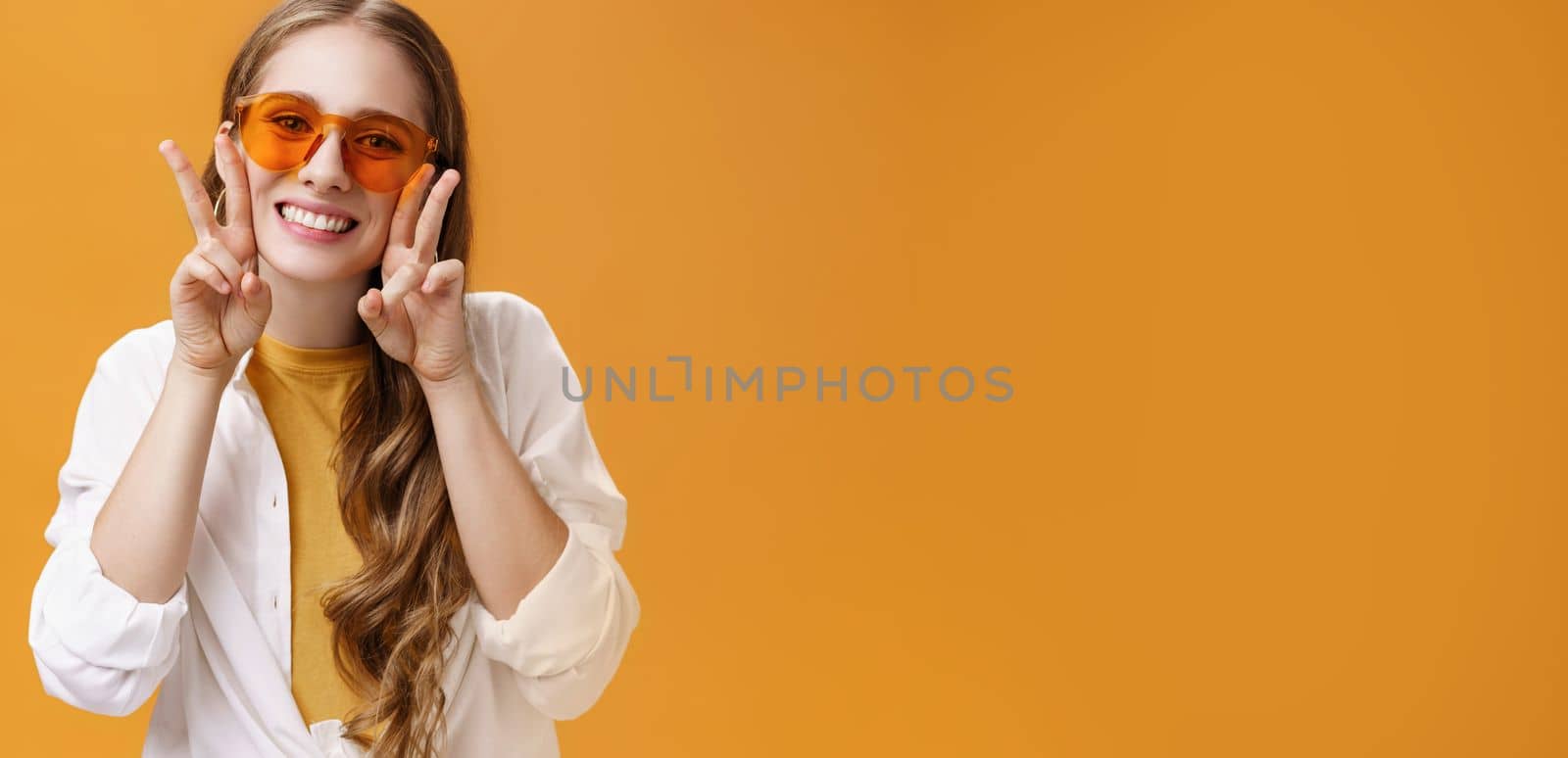 Stylish enthusiastic and charismatic young party girl in trendy sunglasses and white blouse over t-shirt showing peace gestures near face and smiling cute at camera against orange background by Benzoix