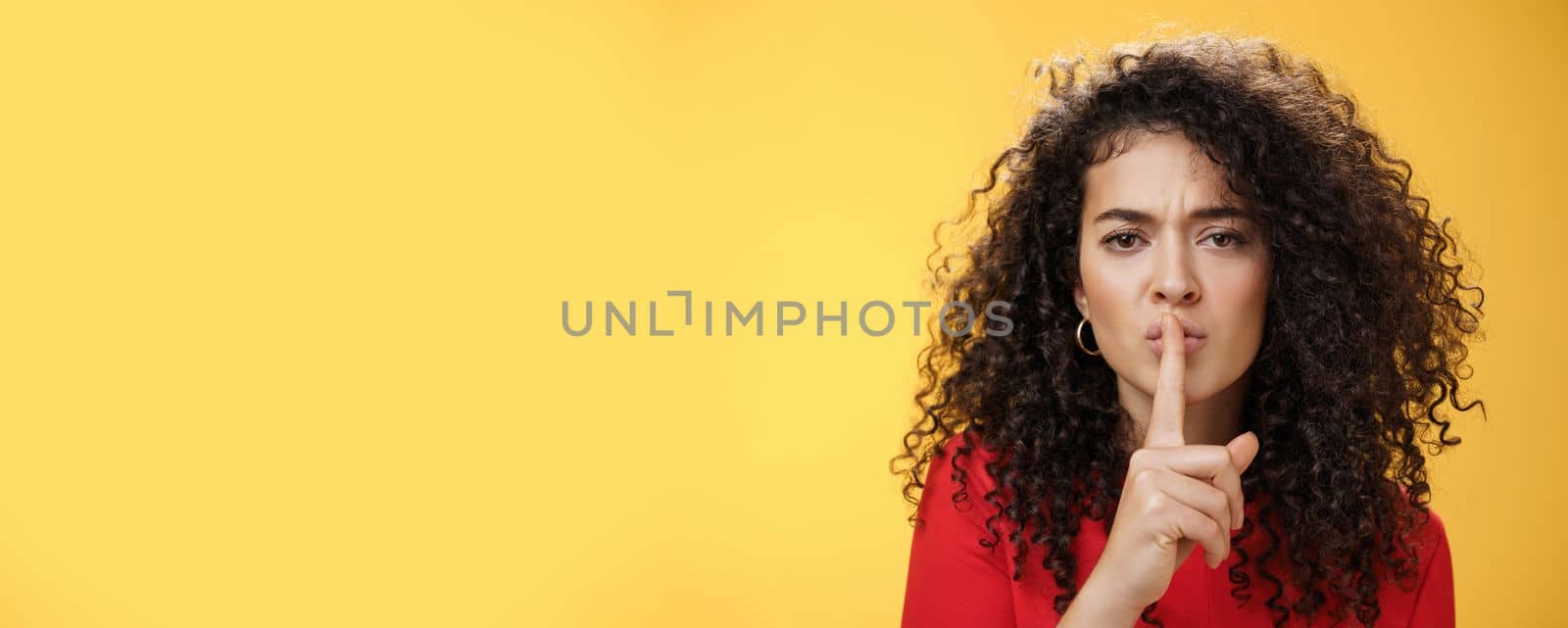 Shh not say word. Portrait of serious-looking sexy young woman with curly hair showing shush gesture with index finger over folded lips frowning asking keep secret promise keep silent over yellow wall by Benzoix