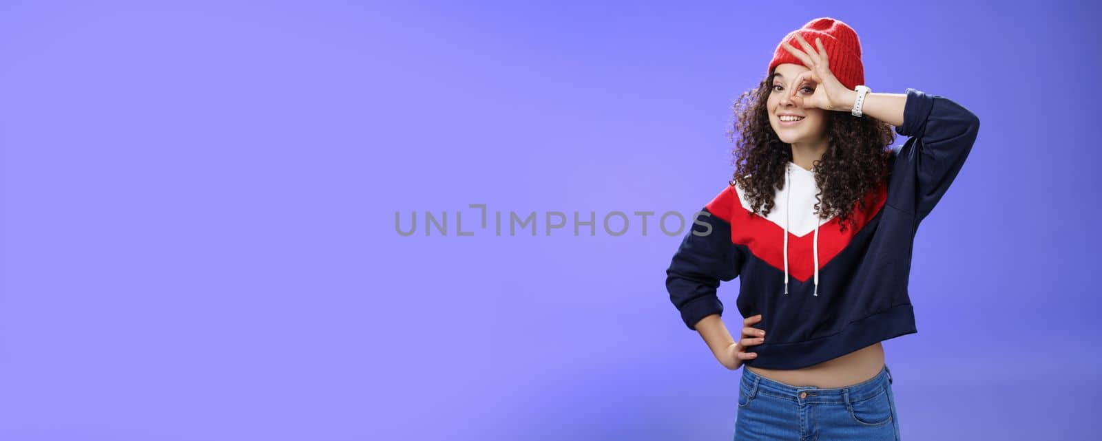 Okay girl agrees. Friendly-looking happy and charming young curly-haired girl in warm beanie showing ok gesture or circle over eye as peeking at camera and smiling broadly with joy over blue wall.