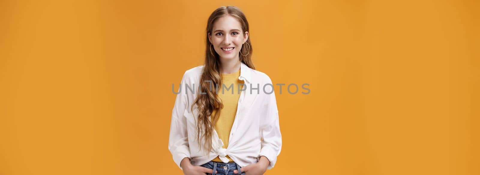Stylish young female designer wants help friend shopping holding hand in pockets smiling joyfully and self-assured at camera wearing trendy blouse over yellow t-shirt posing over orange wall by Benzoix
