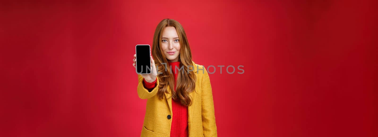 Attractive and stylish ginger girl with confident glance and self-assured smile pulling mobile phone at camera showing smartphone screen grinning delighted as posing against red background by Benzoix