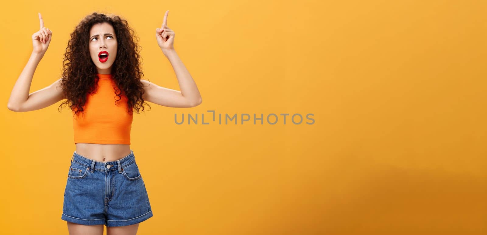 Silly and moody european party girl with curly hairstyle. in red lipstic and stylish orange cropped top complaining on bad weather raising hands pointing and looking up with displeased unhappy face.