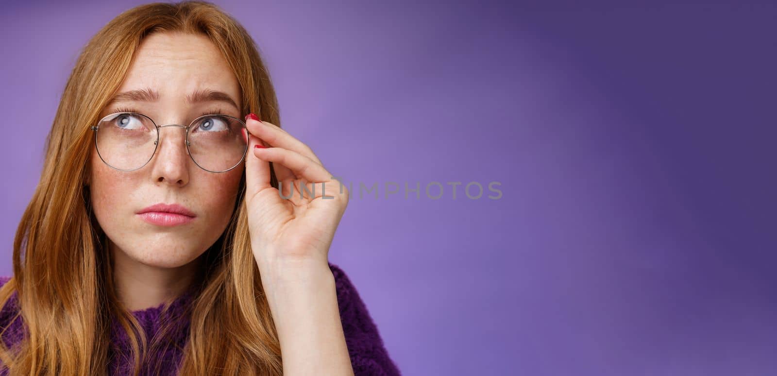 Lifestyle. Clumsy and cute young female teacher in glasses with red hair looking unsure and confused looking interested at upper left corner touching rim of glasses thinking or picturing in imagination.