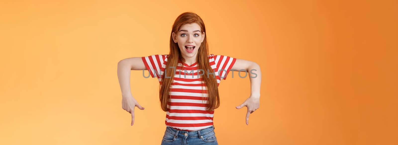 Enthusiastic lively cute redhead woman introduce incredible offer, pointing down, bottom copy space, smiling amazed, look camera, explain you awesome opportunity, feel excitement, orange background.