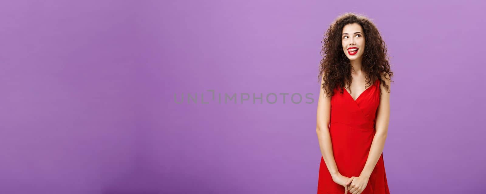 Studio shot of cute thoughtful and creative charming european woman in stylish red licking lips with desire and amusement looking at upper right corner smiling, daydreaming using imagination. Copy space