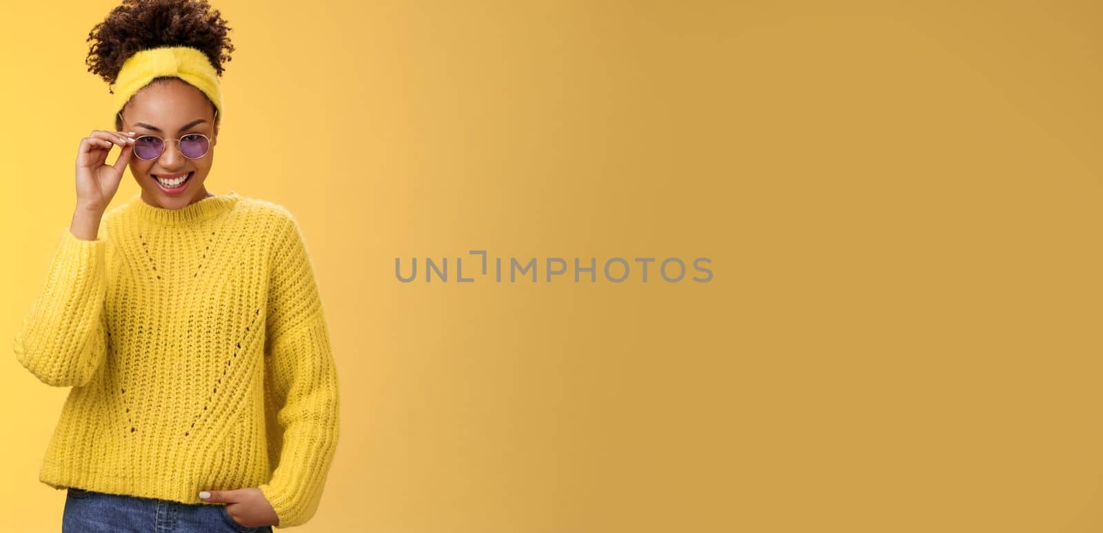 Stylish flirty modern african-american woman afro hairstyle in sweater headband touching sunglasses smiling joyful sparkling joy amazement happy attend awesome fashion party, yellow background.