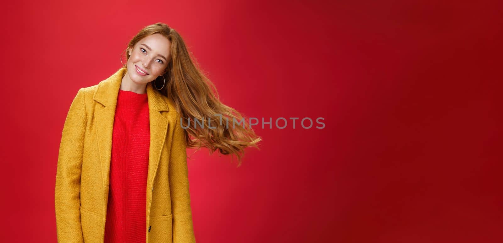 Charming redhead female with freckles waving hair. smiling broadly tilting head as haircut flying in air posing delighted and carefree over red background in warm yellow coat and knitted dress.