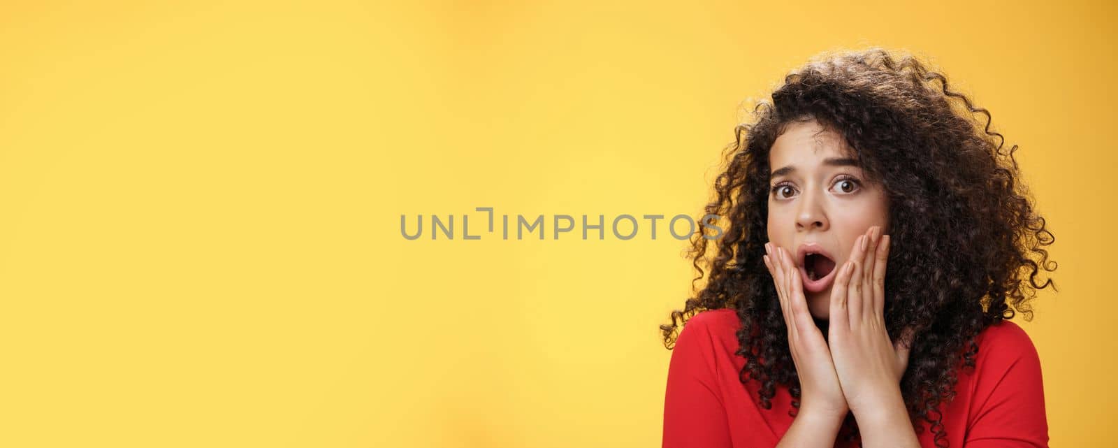 Indoor shot of shocked female drama queen with curly hair gasping in surprise, feeling empathy and fear turning head holding hands on cheeks astonished as hearing shook stunning news. Emotions concept