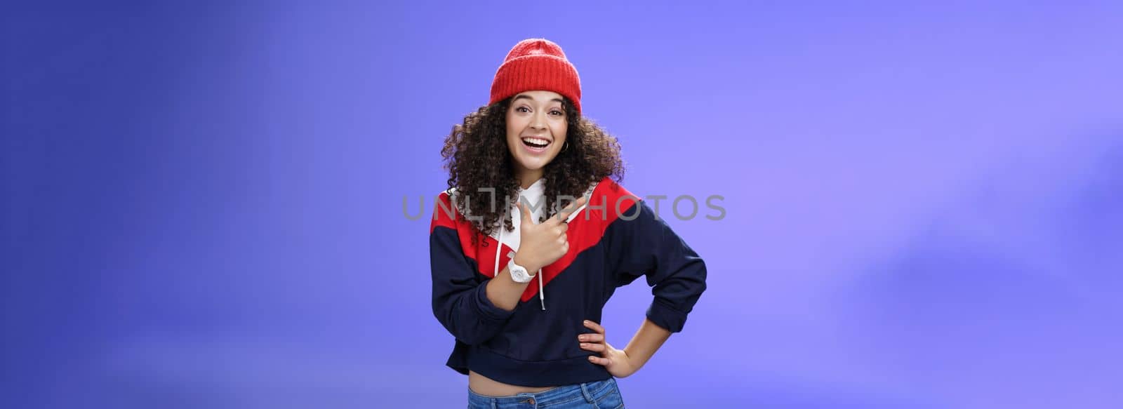 Stylish and sociable friendly-looking attractive woman with curly hair in warm beanie pointing at upper left corner laughing from joy and amazement having fun posing happily over blue background by Benzoix