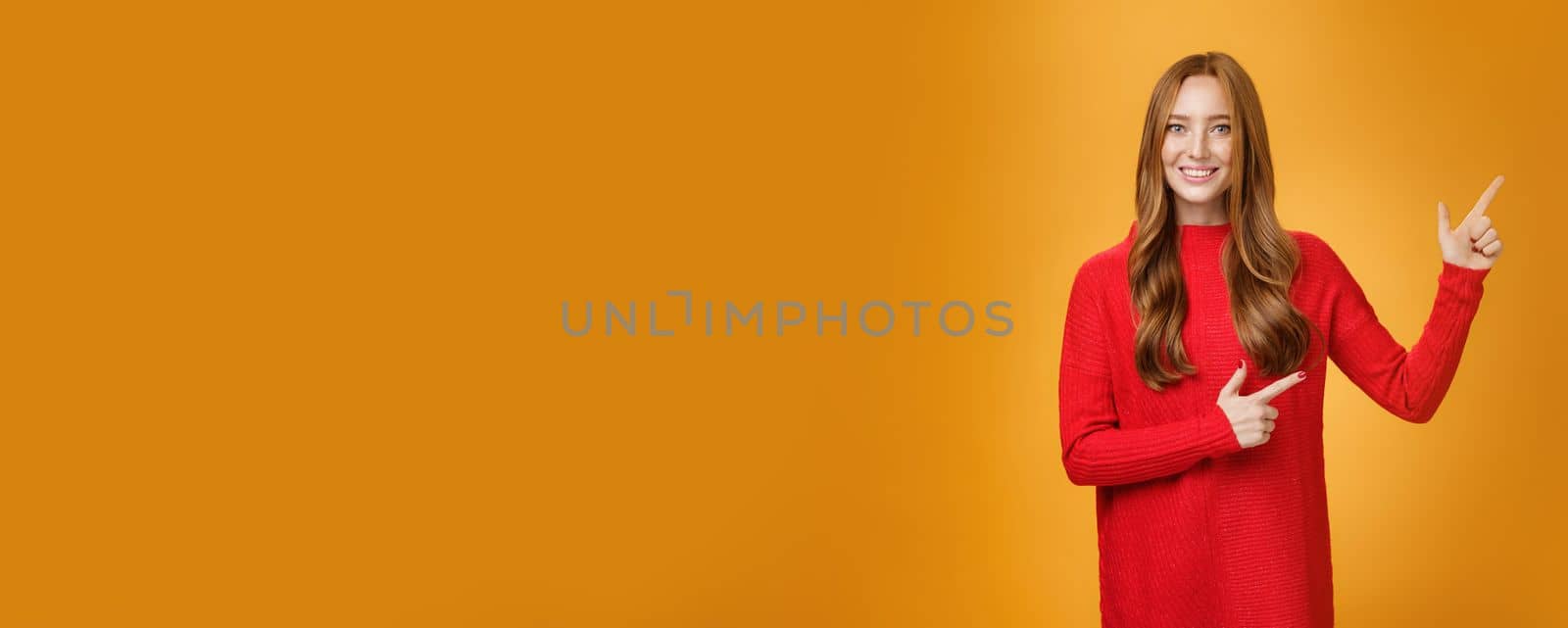 Friendly-looking joyfuly and energized redhead female in red sweater pointing at upper right corner promoting advertisement with broad delighted and tender smile posing against orange background by Benzoix