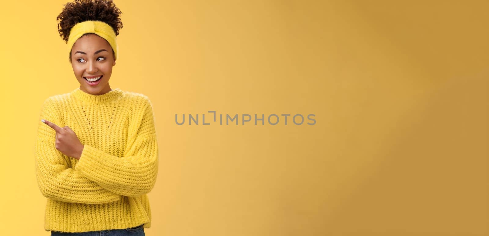 Amused young cute millennial african woman peeking silly pointing left smiling broadly show curious interesting place hang our friends promote product indicating advertisement, yellow background.