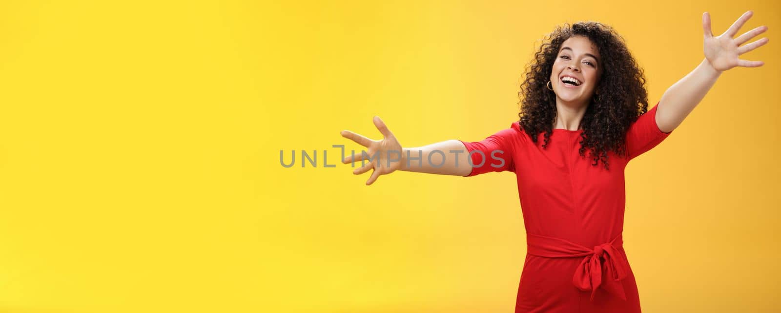 Come into my arms. Portrait of friendly and loving, caring charming woman with curly hair in red casual dress spread hands as wanting give hug smiling broadly at camera giving warm welcome or cuddle by Benzoix