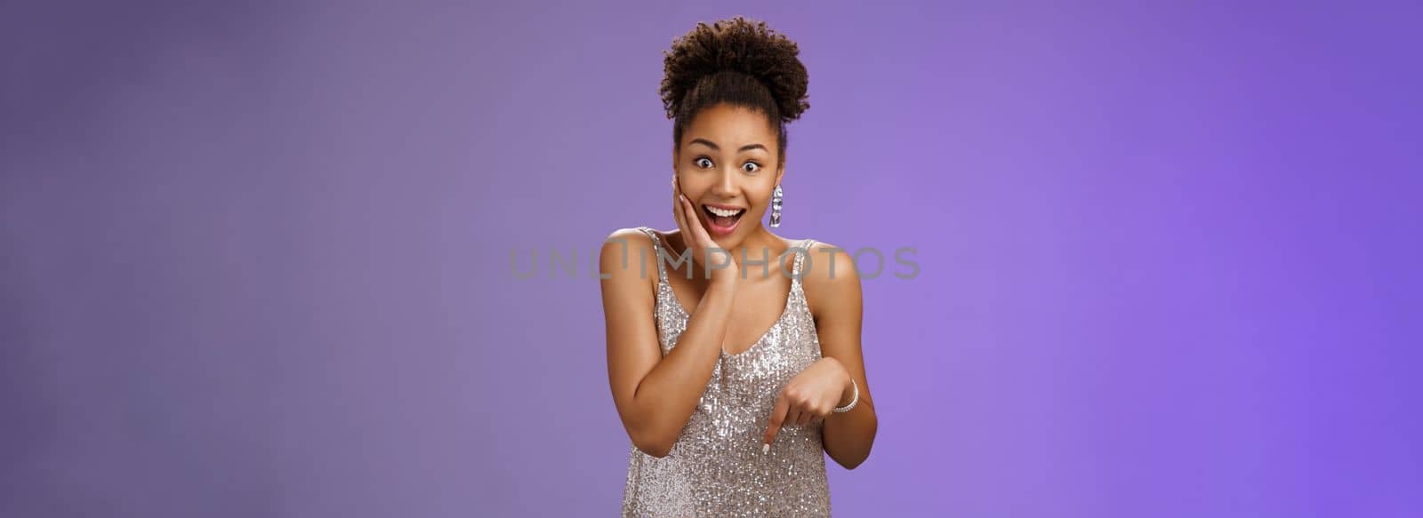 Attractive surprised charismatic african-american woman in silver glittering stylish dress widen eyes gasping amazed touch cheek impressed astonishing perfect gift pointing down cannot believe by Benzoix