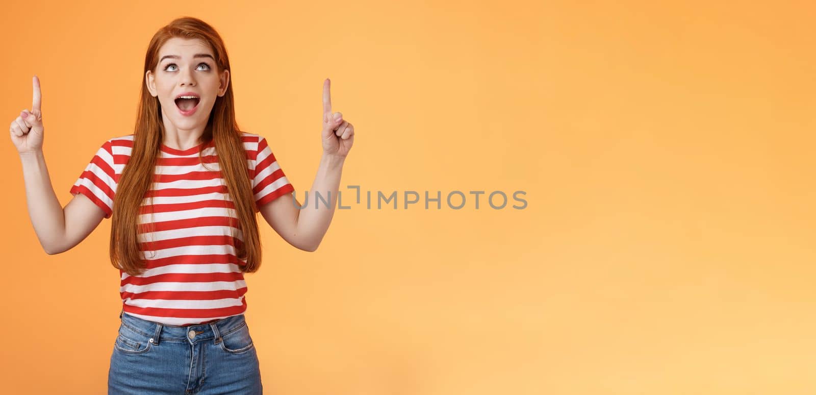 Surprised excited cheerful ginger girl see amazing opportunity, look pointing up amused, drop jaw gasping astonished, pleased awesome cool advertisement, reacting thrilled top copy space.
