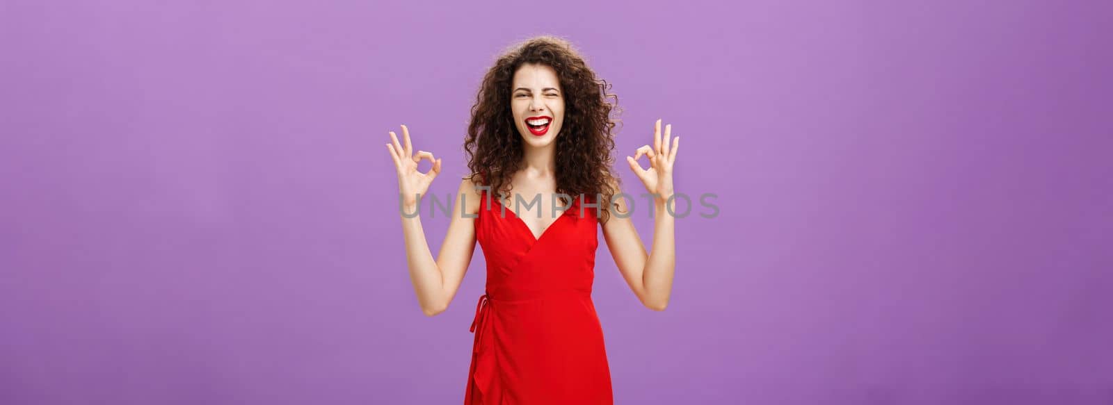 Happy elegant woman hosting party. being assured everything be fine showing okay or excellent gesture with fingers winking and smiling joyfully standing in evening red dress wearing luxurious makeup.