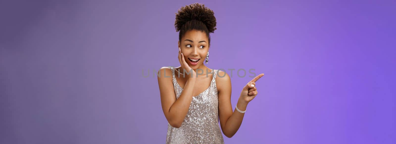 Impressed speechless happy african-american young girl party night club wearing glittering evening dress gasping amused pointing left astonished touch face see famous rock-star, blue background.
