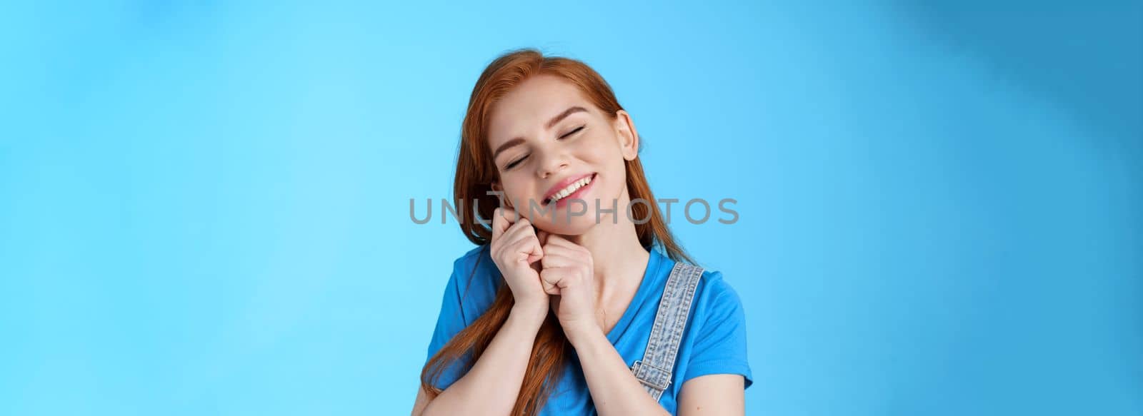 Glad cute tender redhead female, tilt head dreamy lean palms, close eyes and smiling lovely, daydreaming, recall nice memories, cherish moments, stand silly blue background nostalgic mood.