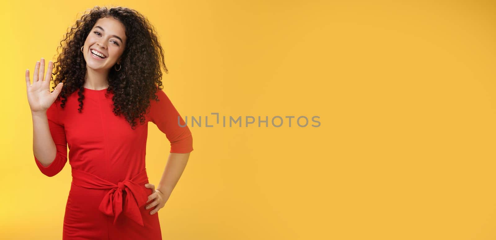 Hey my name is. Friendly-looking self-assured carefree cute 25s woman with curly hair waving with raised palm in hello or hi gesture smiling broadly greeting new coworkers over yellow background.