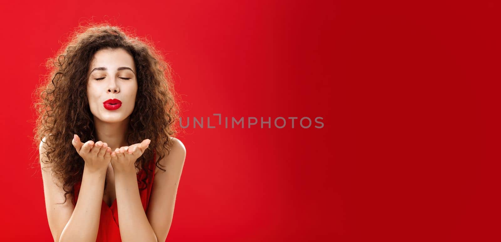 Tender and gentle stylish caucasian girl with curly hairstyle and red lipstick bending towards camera with slight smile closed eyes and palms near folded lips blowing kiss at camera romantically by Benzoix