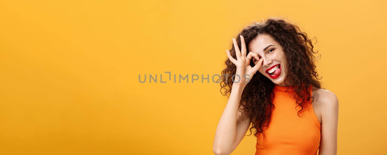 Carefree optimistic and energized good-looking woman with curly hairstyle tilting head and sticking out tongue playfully showing okay or excellent sign over eye posing near orange background by Benzoix