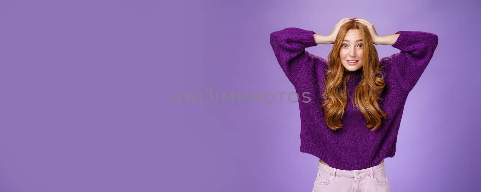 Cute european redhead 20s woman acting nervous, holding hands on head raising eyebrows in worry and anxiety, panicking being troubled think up plan posing over purple background. Copy space