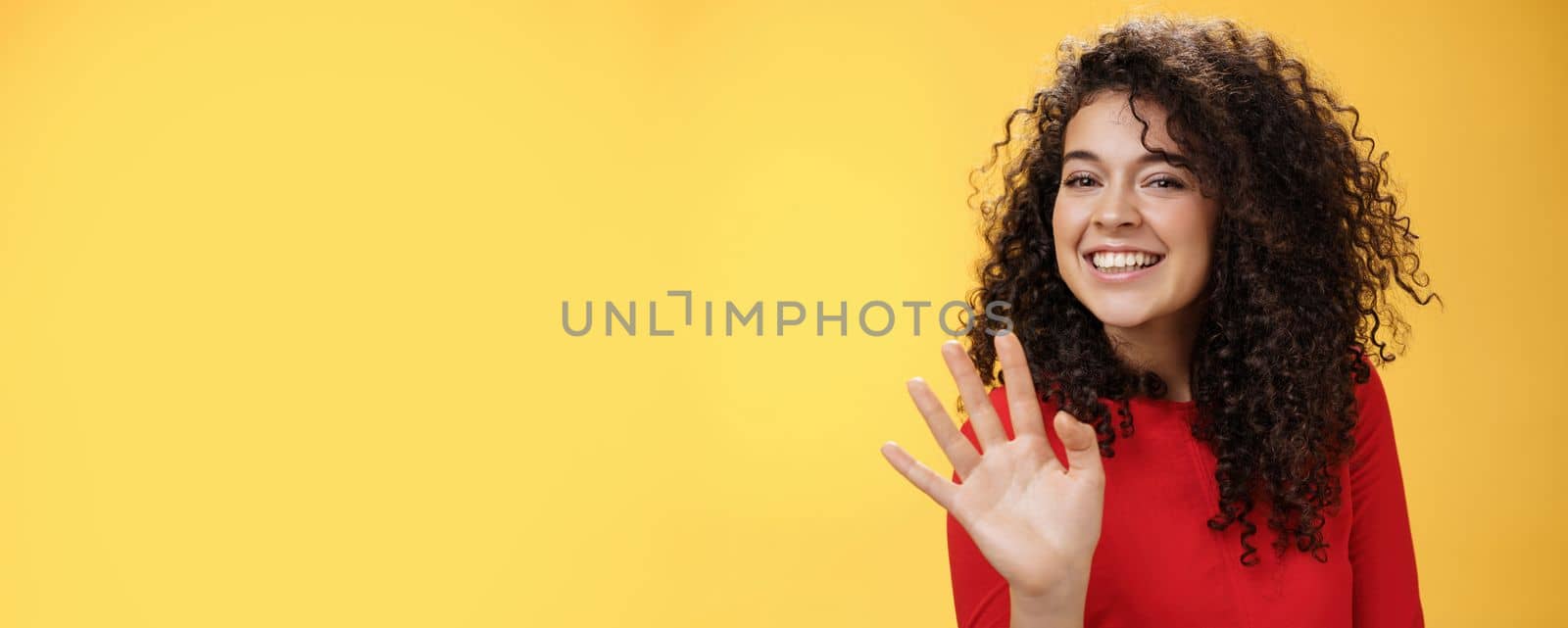 Lifestyle. Charming friendly and self-assured attractive curly woman waving cute with palm to say hi or hello smiling broadly greeting man trying flirt in party posing joyful over yellow background.