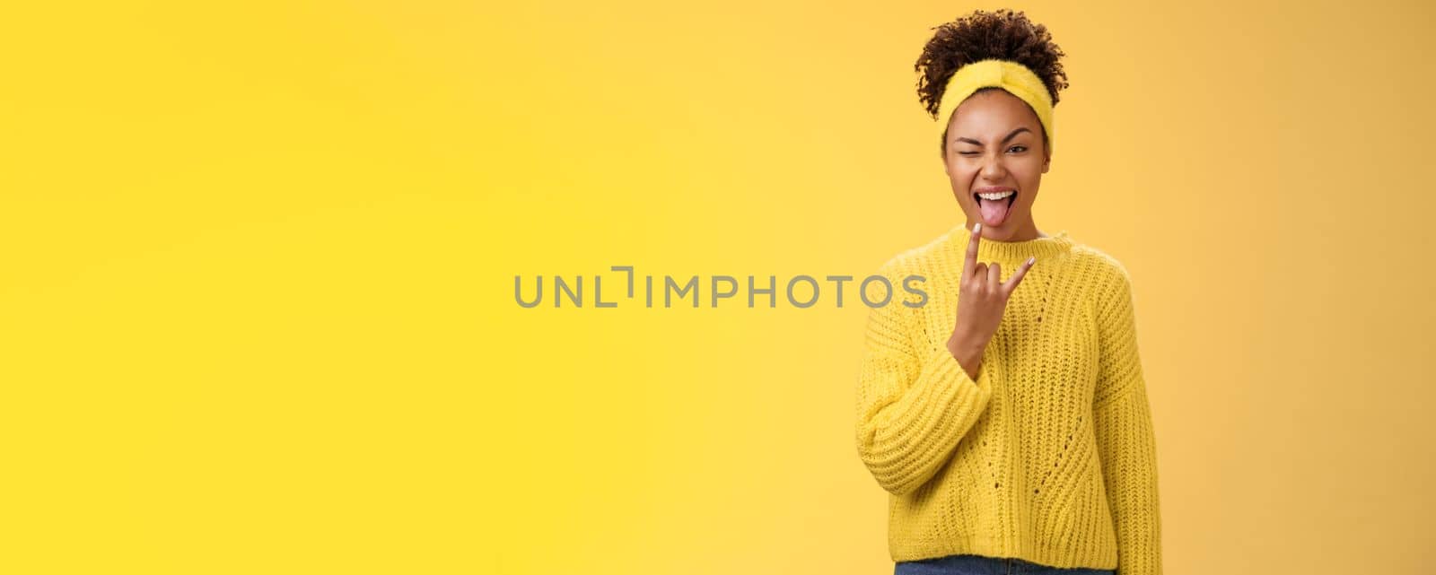 Excited charming smiling daring sassy young african american woman having fun show tongue rock-n-roll heavy metal gesture winking happily enjoying cool awesome party, standing yellow background.