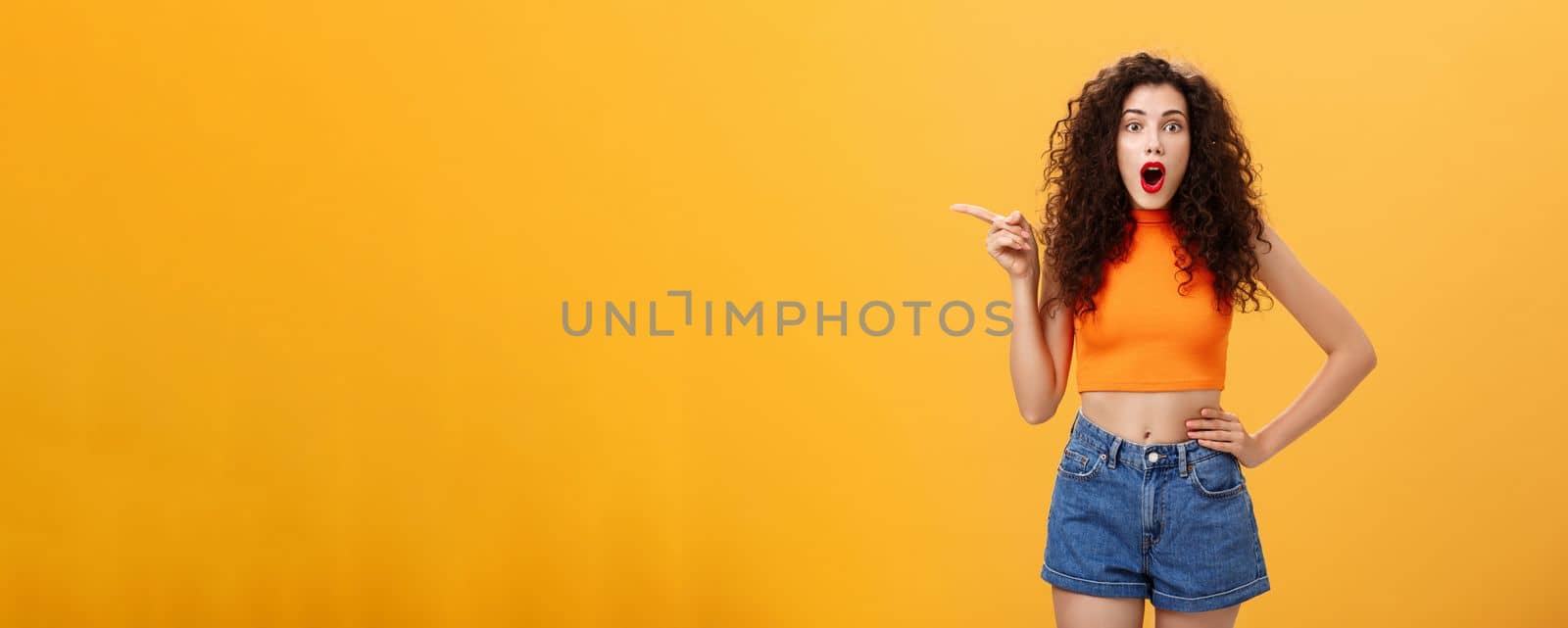 Silly and moody european party girl with curly hairstyle. in red lipstic and stylish orange cropped top complaining on bad weather raising hands pointing and looking up with displeased unhappy face.