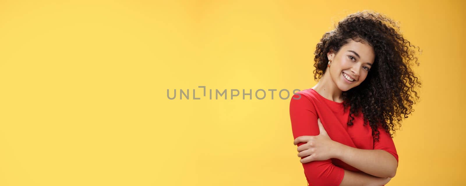 Waist-up shot of tender feminine and gentle woman with curly hairstyle combed to right side, tilting head and smiling flirty making romantic gazed at camera hugging herself over yellow background by Benzoix