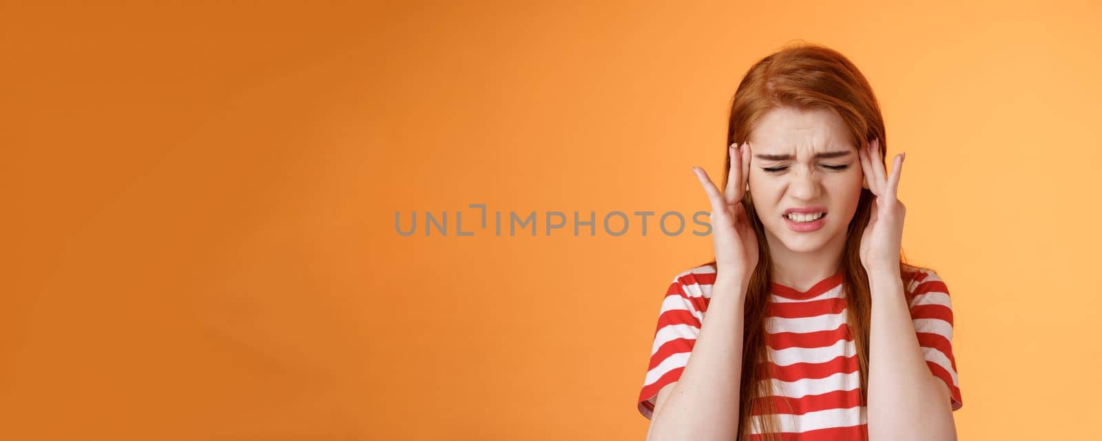 Dizzy uneasy cute redhead female student suffering huge headache, cringe, frowning clench teeth painful feeling, touch temples, feel pain head, awful migraine, asking painkillers, orange background.