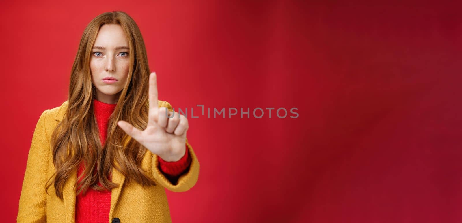 Serious-looking redhead woman in yellow coat extending hand to camera with stop and warning gesture prohibiting and forbidding make something having unemotional face as being confident over red wall by Benzoix