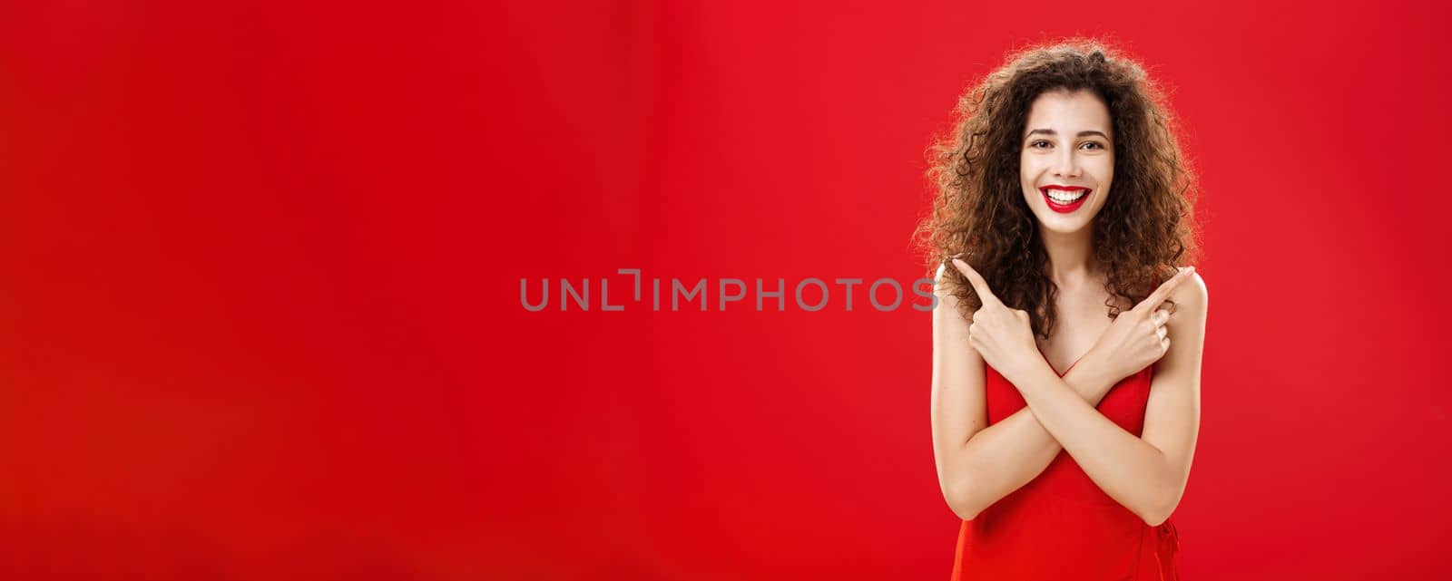 Woman satisfied with any decision feeling carefree and happy standing in elegant red evening dress with curly hairstyle crossing hands on chest pointing left and right delighted with choices. Copy space