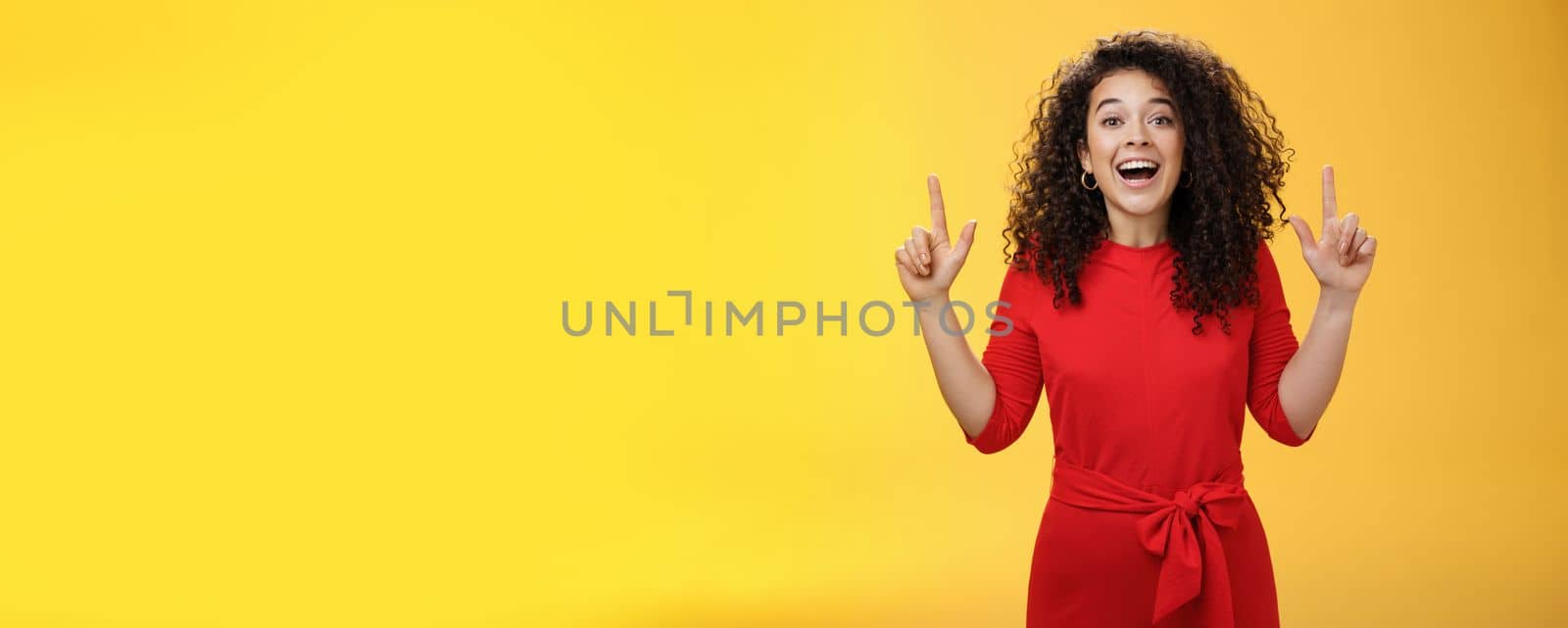Enthusiastic happy curly-haired young european woman feeling happy present awesome copy space, raising hands pointing up and smiling with delight and admiration over yellow background.