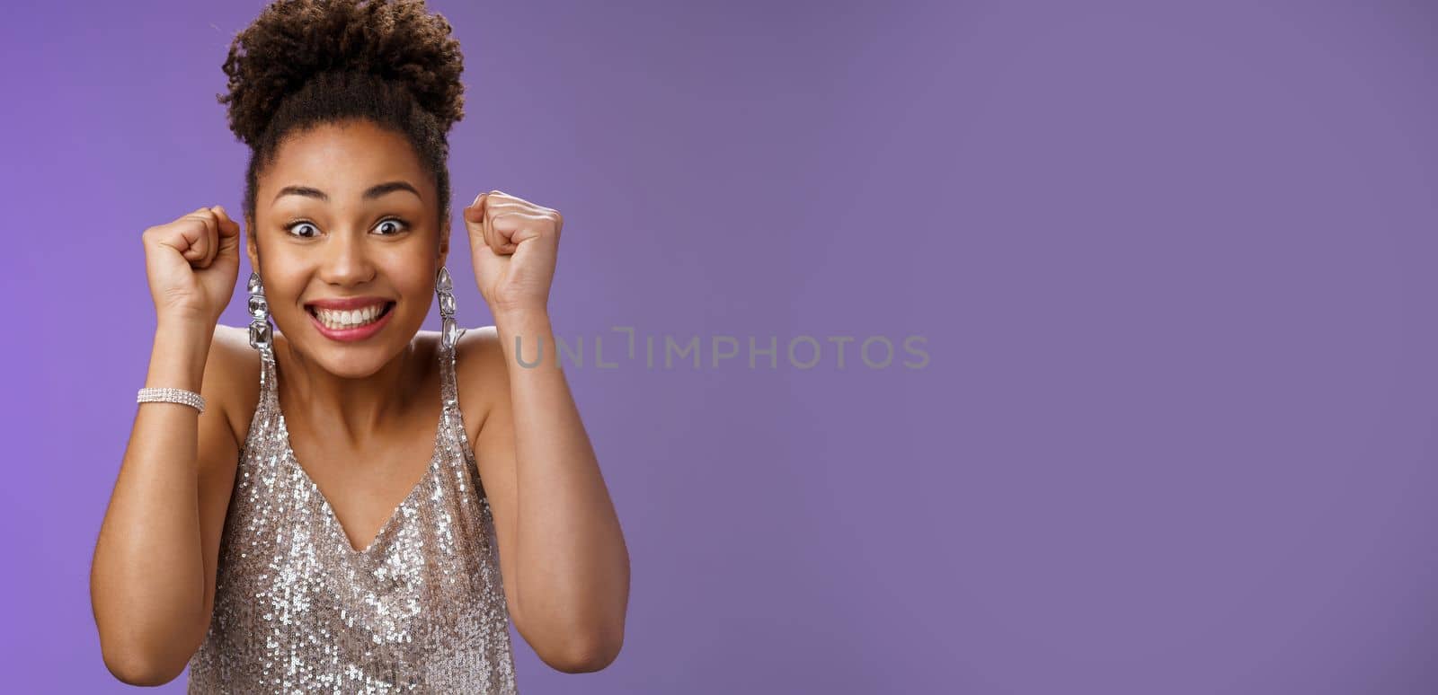 Excited african woman in elegant silver dress brilliants smiling thrilled winning clench raised fists grinning triumphing rejoicing receive perfect news celebrating, standing blue background.