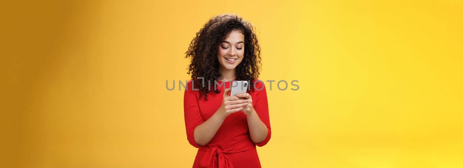 Lifestyle. Gil sending message spread news across social network having party inviting friends via smartphone holding mobile phone in hands smiling broadly at device screen as posing over yellow background.