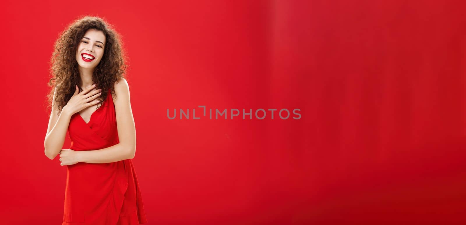 Portrait of elegant pleased and grateful charming caucasian woman with curly hairstyle holding palm on chest smiling and laughing amused posing in elegant evening dress over red background by Benzoix