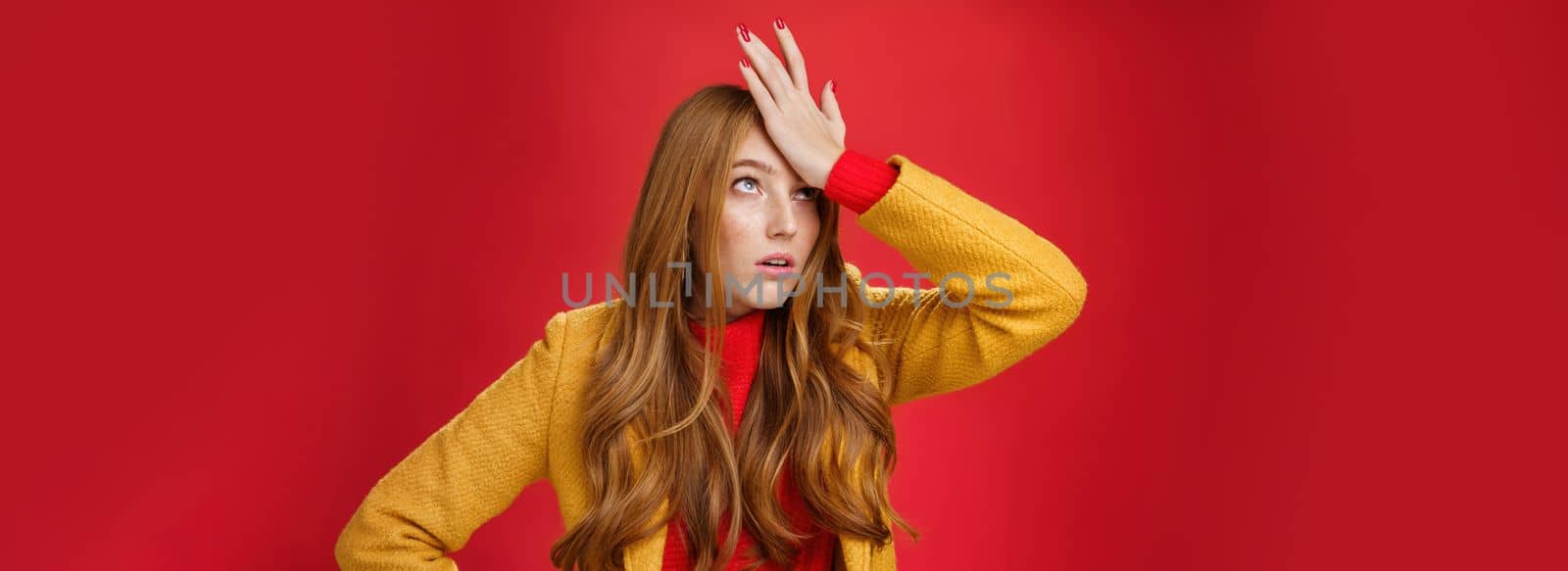 Redhead businesswoman making facepalm gesture with hand on forehead rolling eyes up from annoyance and irritation as being shocked with how dumb client is sighing bothered over red wall, tired.