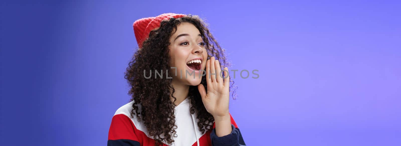 Friend shouting through street to turn attention. Portrait of carefree happy cute woman in beanie and sweatshirt holding palm near opened mouth looking with smile left as calling mate with loud yell.